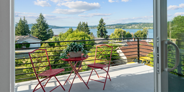 Man sieht eine geöffnete Balkontür, die auf einen Balkon mit einem roten Tisch und zwei roten Stühlen führt. Im Hintegrund sieht man einen großen See