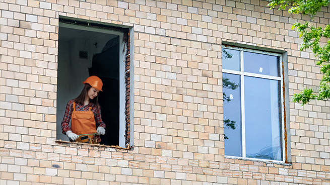 Ein neues Fenster und eine leere Fensteraussparung in einer Hauswand. Eine Frau misst gerade die Fensterbreite.