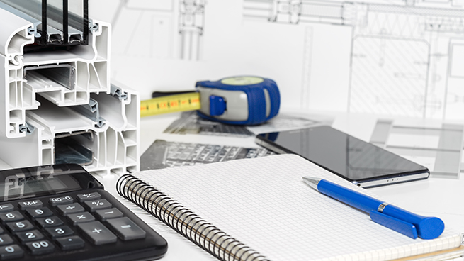 Schreibtisch mit Maßband, Schreibblock und Stift und Musterecke. Im Hintergrund sieht man technische Zeichnungen und Grundrisszeichnungen