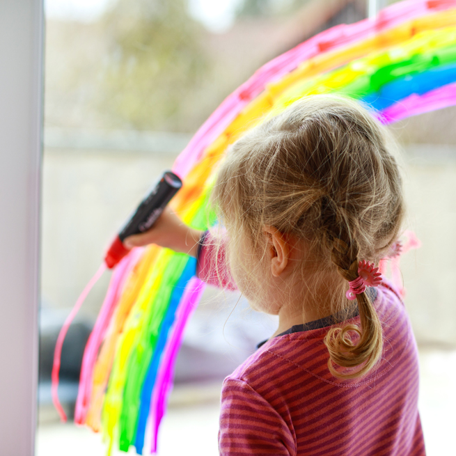 Mädchen malt mit Fensterfarben einen Regenbogen in ein Wohnraumfenster