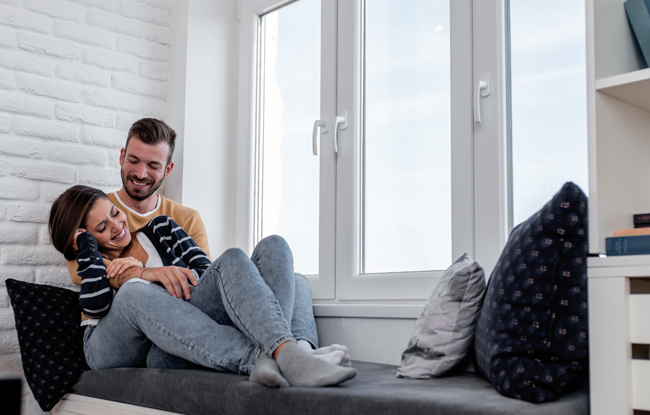 Dreiflügeliges, weißes Kunststofffenster mit Sitzbank davor, auf der ein Mann und eine Frau zusammen sitzen und lachen