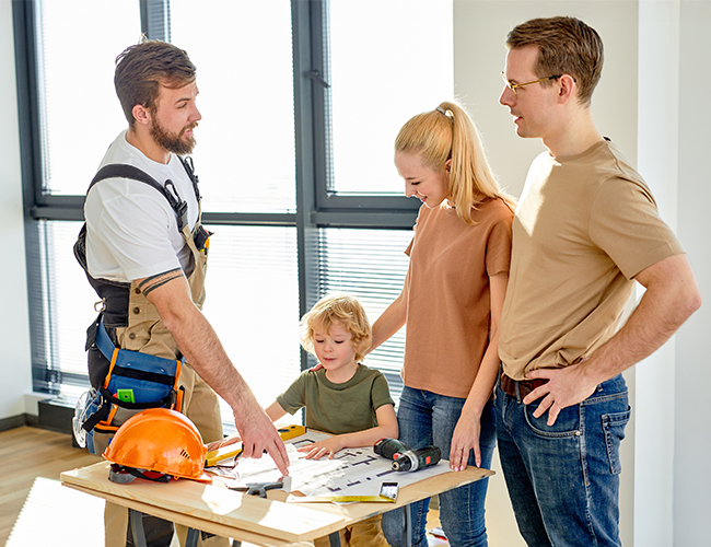 Eine Familie steht mit einem Handwerker an einem Tisch und schaut sich Baupläne an