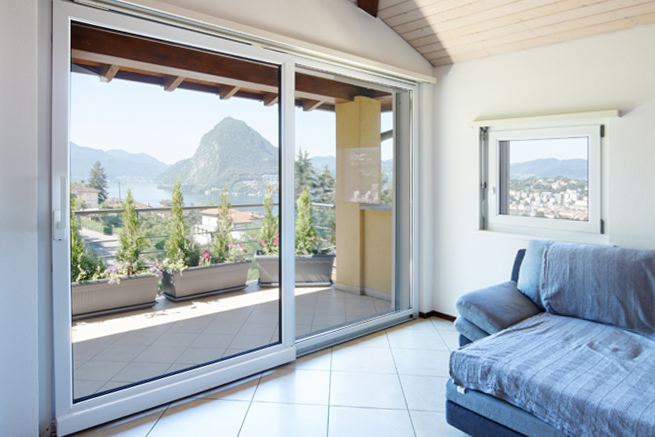 Zimmer mit Aussicht durch eine Parallel-Schiebe-Kipp-Türe auf einen Balkon mit Blick auf eine Stadt am See