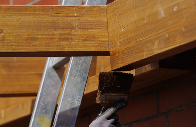 Holzbalken einer Terrassenüberdachung werden auf einer Leiter stehend gestrichen