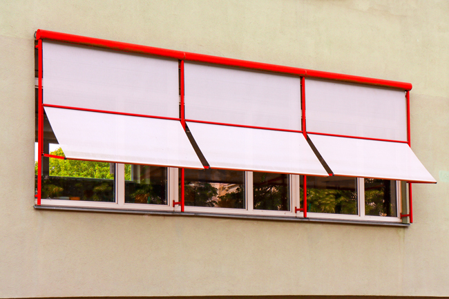 Drei helle Fenstermarkisen in roten Gestellen vor einem dreiteiligen Fenster